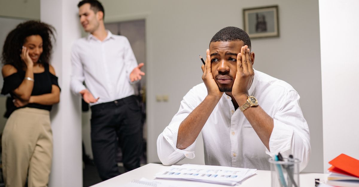 A stressed employee sits at a desk with colleagues in discussion nearby, highlighting workplace tension.