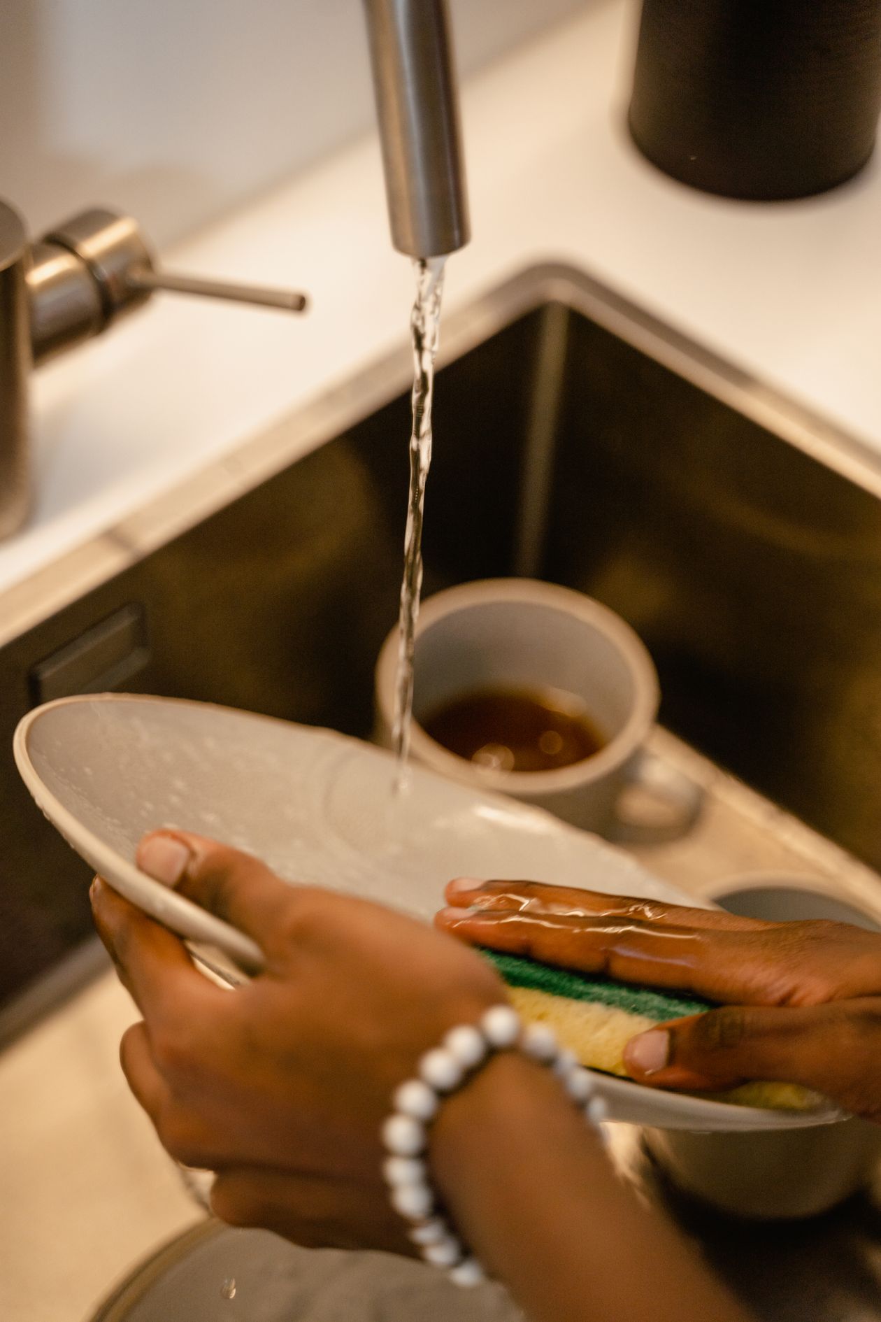 A person washing dishes with a sponge under a flowing faucet in a kitchen sink