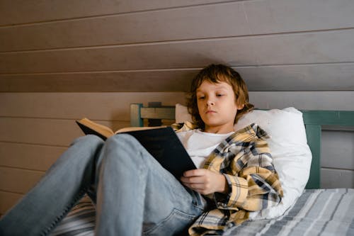 A Boy Holding a Book While Lying on the Bed Near the Wooden Wall