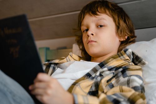 Free A young boy comfortably lying on a bed in his room, deeply engrossed in reading a book. Stock Photo