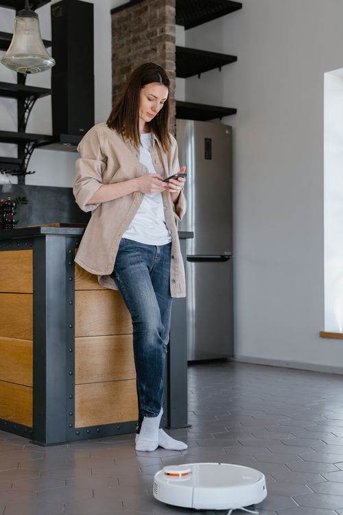 Free Woman controlling robot vacuum with smartphone in a modern kitchen setting. Stock Photo