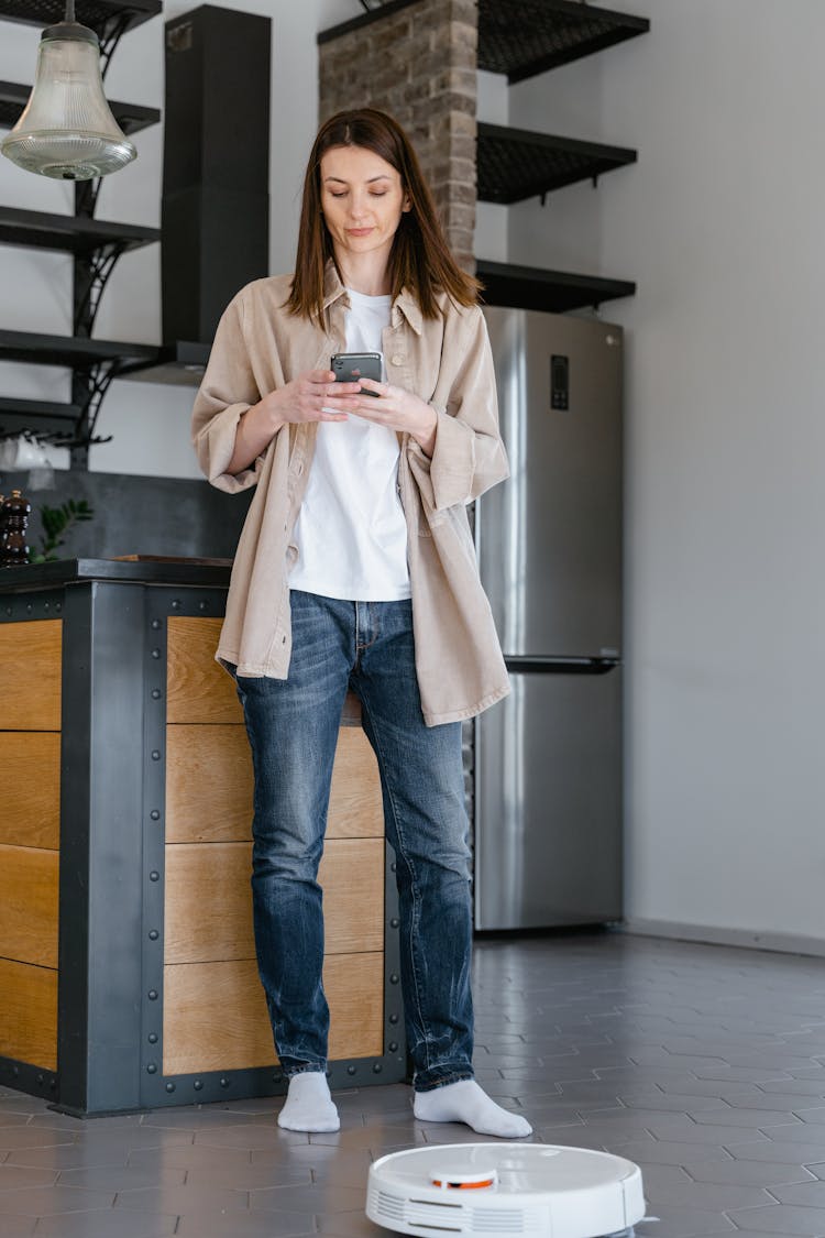 Woman In Beige Long Sleeve Shirt And Blue Denim Jeans Standing Beside Kitchen Counter While Texting