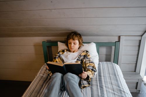 Free A young boy lies on a bed reading a book in a cozy indoor setting, enjoying quiet time. Stock Photo