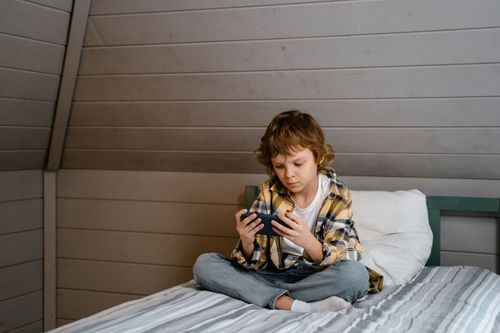 Free Young boy sits on bed focused on a mobile phone, wearing casual attire in a cozy bedroom. Stock Photo