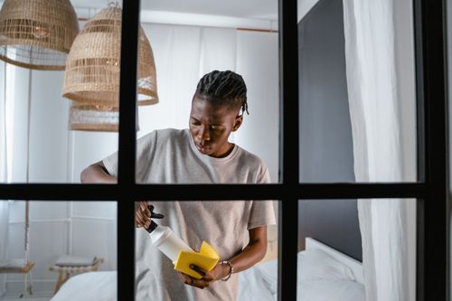 Free A man with dreadlocks cleans a bedroom using a spray bottle and yellow sponge. Stock Photo