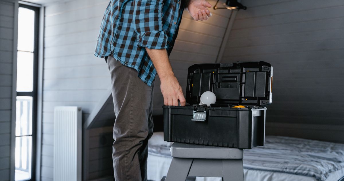 A man in a checked shirt reaching into an open black and yellow toolbox on a step stool in a bright bedroom