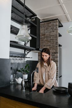 A woman washing dishes at a kitchen sink with stylish interiors and ample lighting.