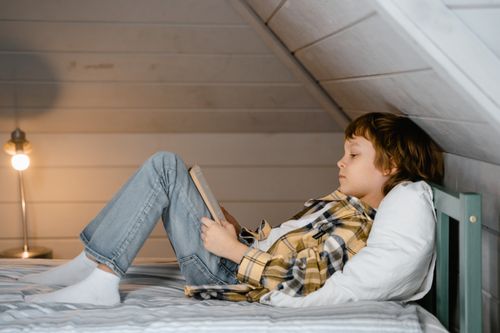 Free A young boy in plaid relaxes with a book in a cozy attic bedroom setting. Stock Photo