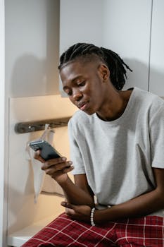 A young man with a smartphone wearing a gray shirt and red plaid pants indoors.