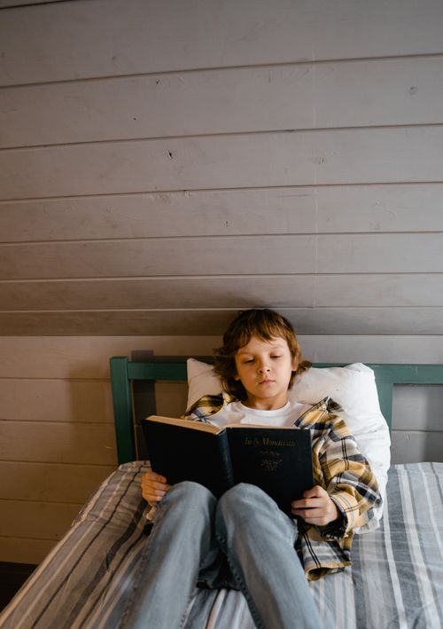 Free A child relaxes on a bed, deeply engrossed in a book, against a wooden wall backdrop. Stock Photo