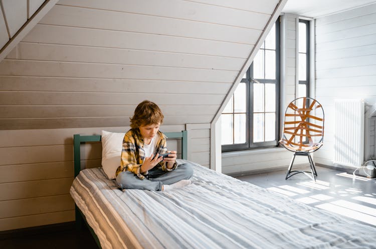 A Kid Sitting On The Bed Using A Mobile Phone