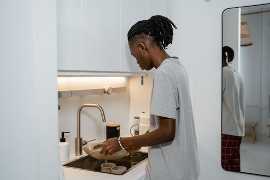 A man washing dishes at the kitchen sink in a modern, well-lit apartment.