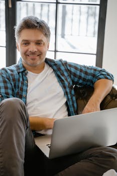 A man in a casual shirt smiles while using a laptop indoors, sitting comfortably.