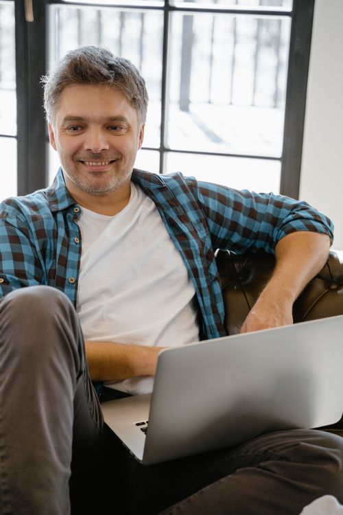 Free A man in a casual shirt smiles while using a laptop indoors, sitting comfortably. Stock Photo