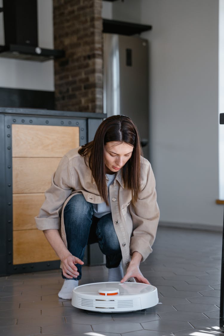 Woman In Brown Coat And Blue Denim Jeans Touching Round Robot Vacuum On The Floor