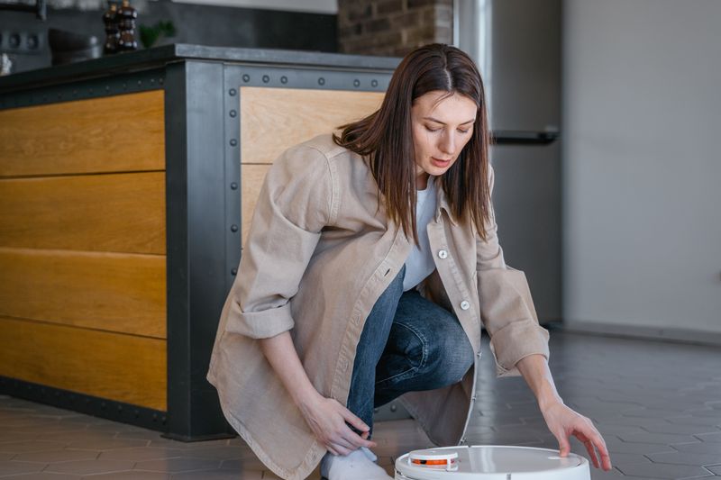 A woman kneeling while managing a robotic vacuum cleaner in a modern kitchen space.
