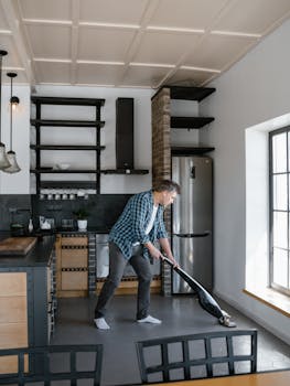 Adult man vacuuming a spacious, modern kitchen with natural light streaming in through large windows.