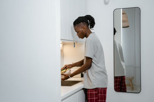 A young man in a gray t-shirt washing dishes in a modern kitchen with a mirror reflection.