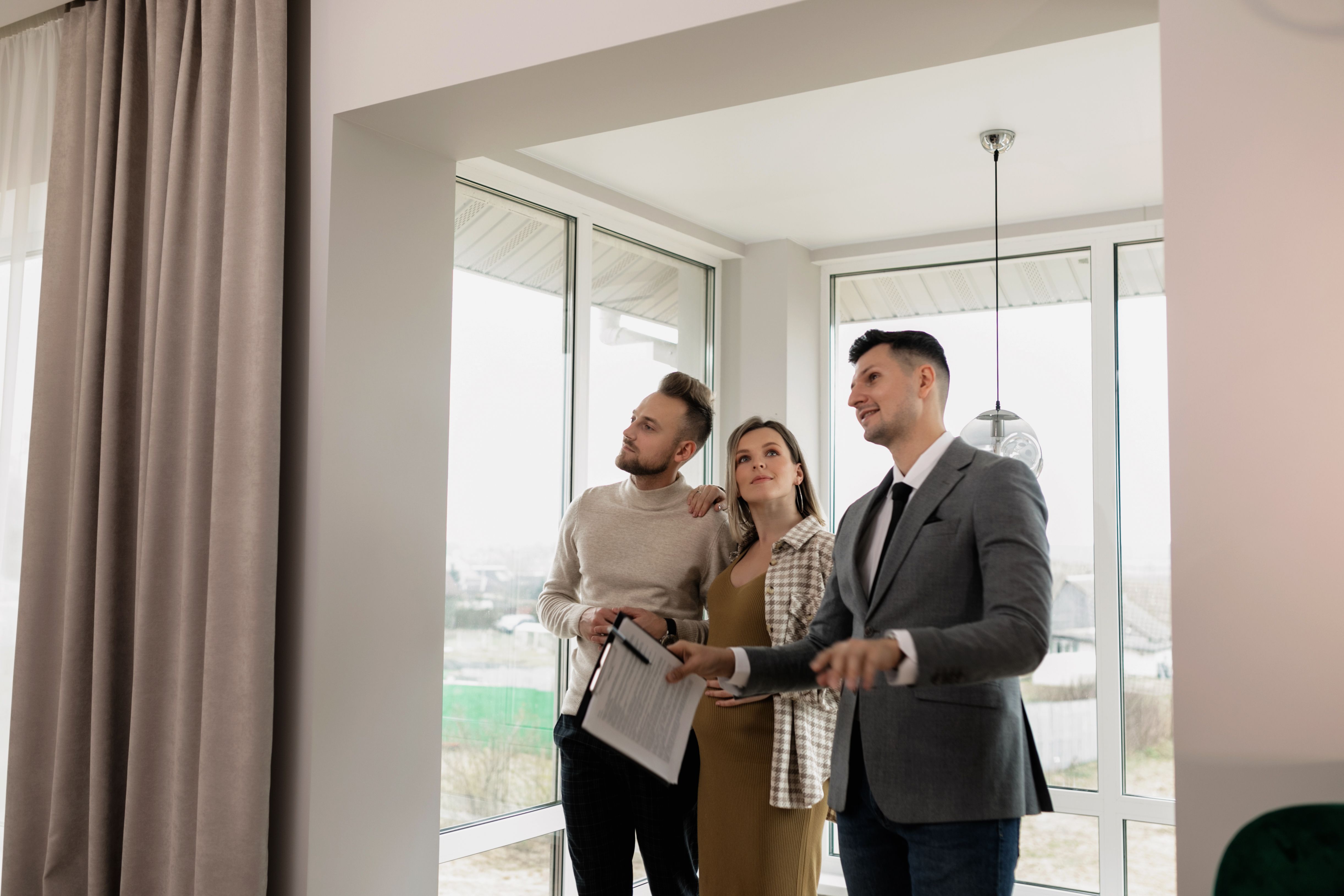 Alt Text: A professional realtor showing a modern house interior to a couple standing near large, bright windows during a private walkthrough.