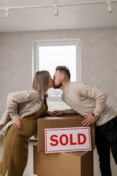 Joyful couple kissing in their new home with 'sold' sign and boxes.