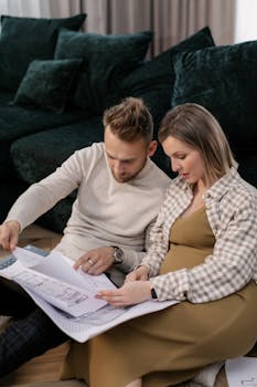 A pregnant couple sits on a floor looking at home blueprints, planning their future living space.