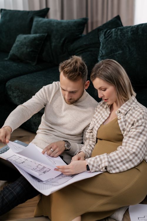 Free A pregnant couple sits on a floor looking at home blueprints, planning their future living space. Stock Photo