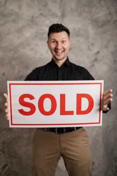 Cheerful real estate agent holding a sold sign, indoors, symbolizing success in property sales.
