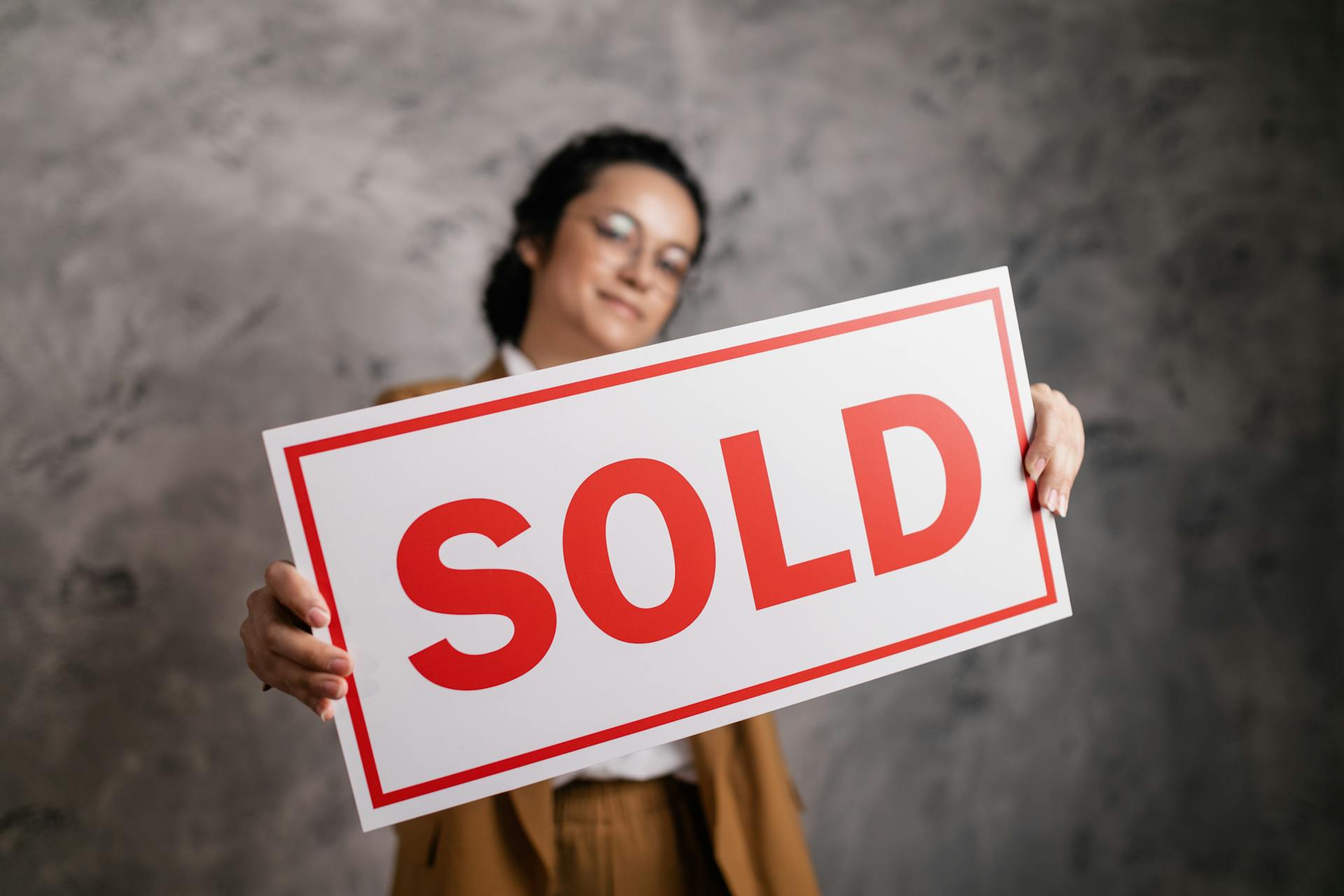 A smiling woman with eyeglasses holding a 'Sold' sign, signifying success in real estate sales.