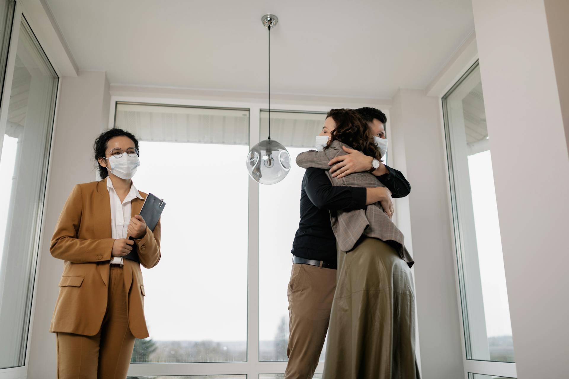 Couple celebrating new home purchase with embracing hug while realtor observes, all wearing face masks.