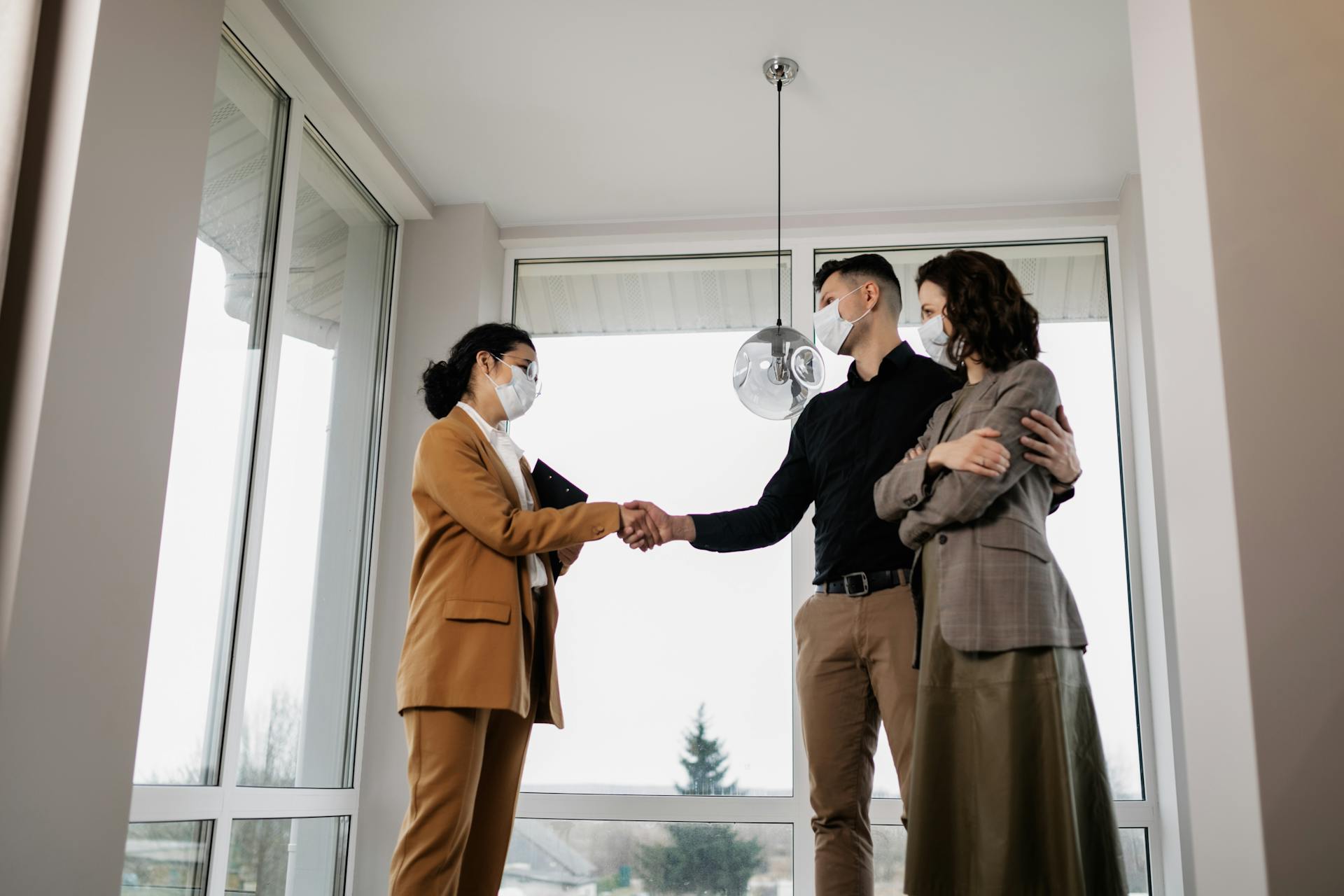 Real estate agent closing deal with couple indoors, all wearing face masks for safety.