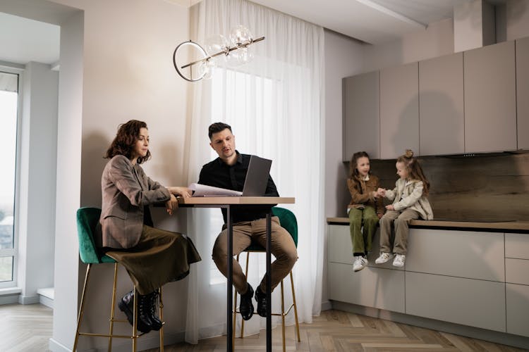 
A Couple Looking At Documents While Their Daughters Are Sitting On A Kitchen Counter