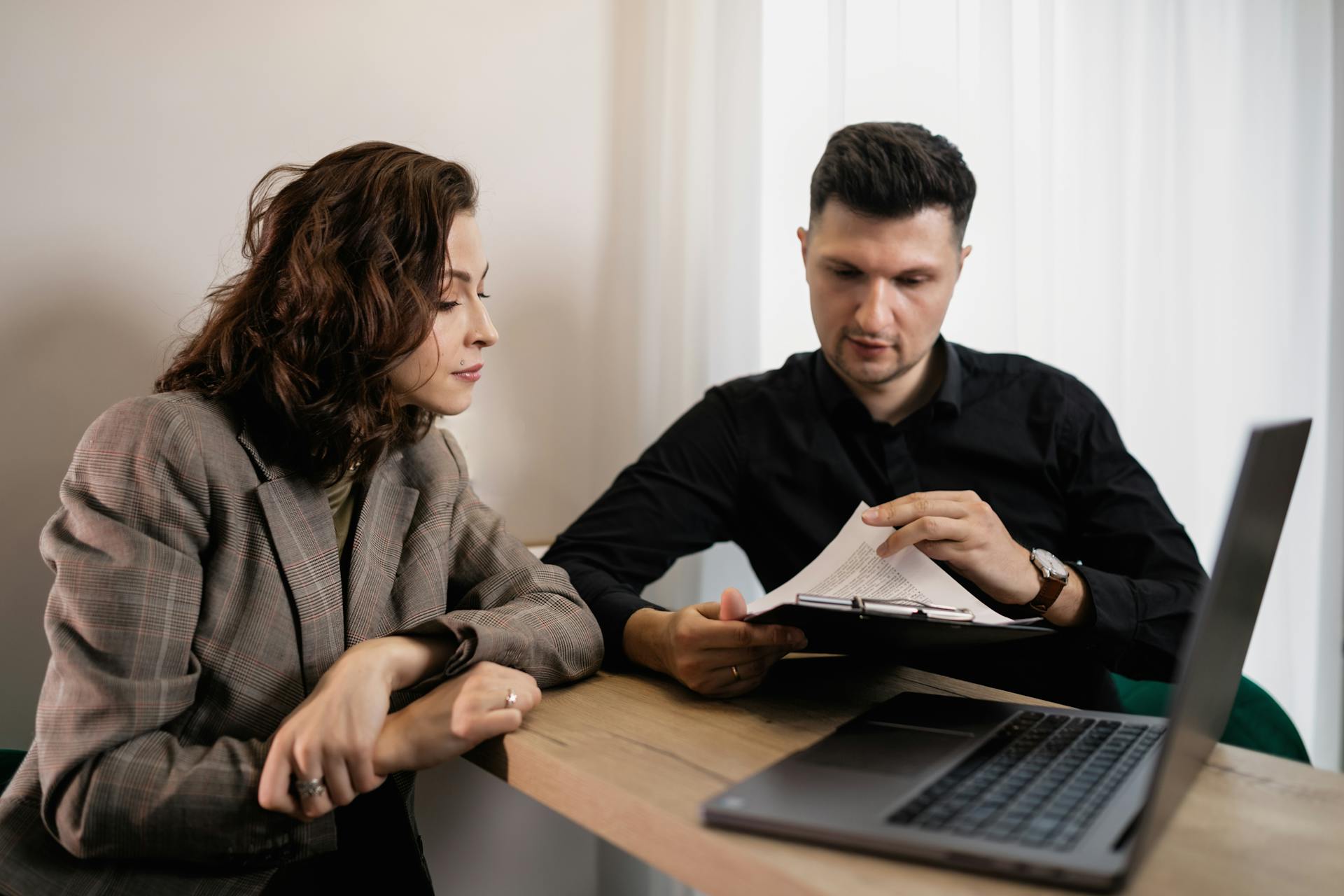 Two professionals discussing documents at a desk with a laptop, focused on collaboration.