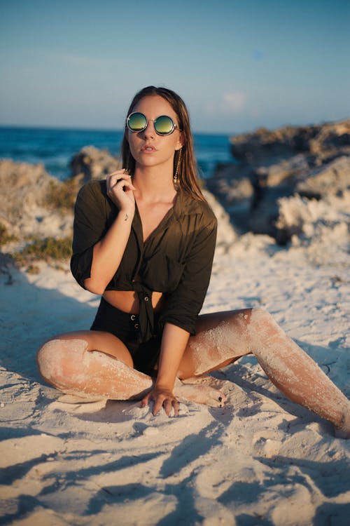 Female in sunglasses and black swimsuit sitting on sandy beach in sunny day