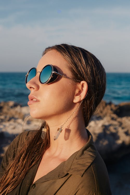 Trendy young woman in earrings and sunglasses on sea shore under cloudy sky in sunlight