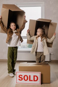 Two girls playfully holding boxes over their heads in a new home with sold sign.