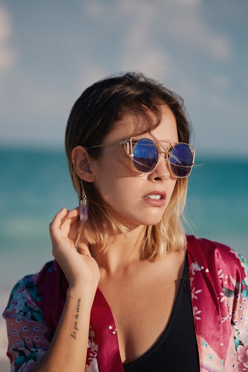 Stylish young tattooed woman in modern sunglasses touching hair on coast in sunlight on blurred background