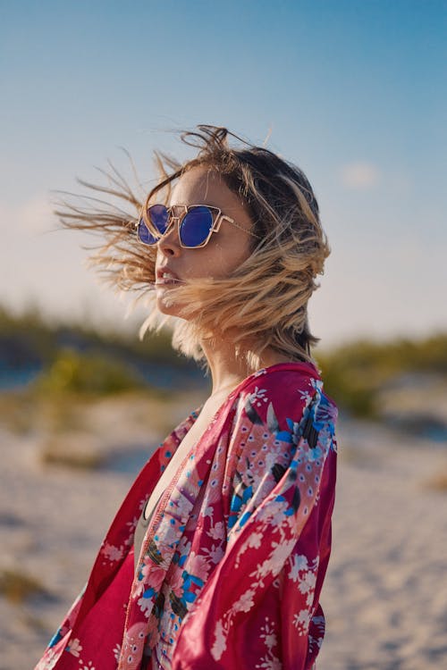 Side view of trendy young female in floral clothes and modern sunglasses looking away on beach in windy weather