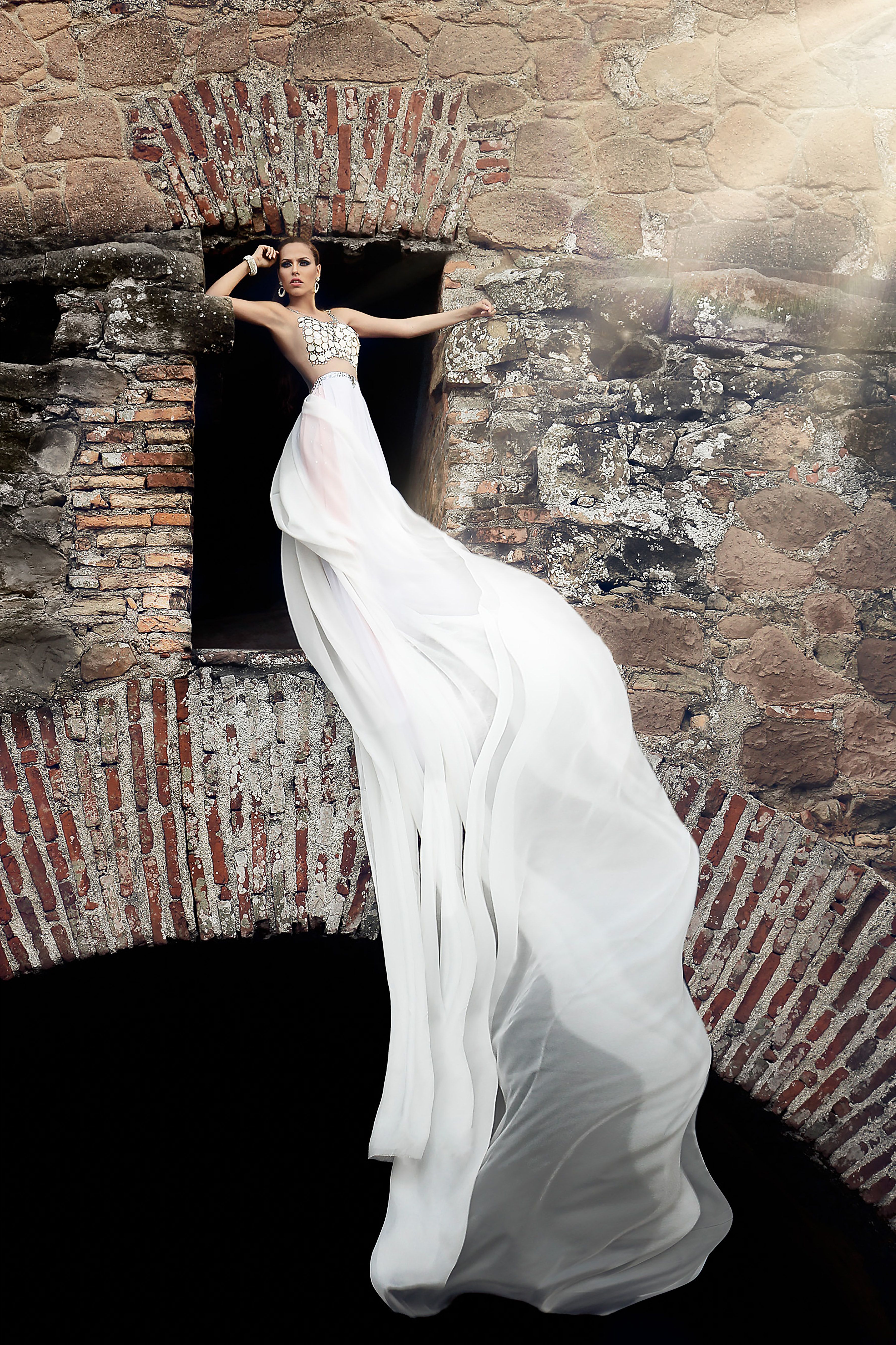 Model in long white dress posing in a historic stone archway, emphasizing elegance and style.