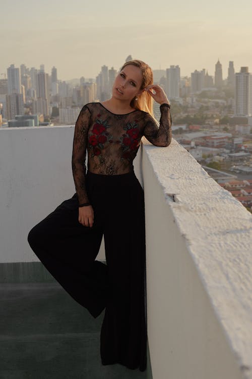 Trendy young woman in transparent lace blouse and black trousers leaning on fence of rooftop while looking at camera in town