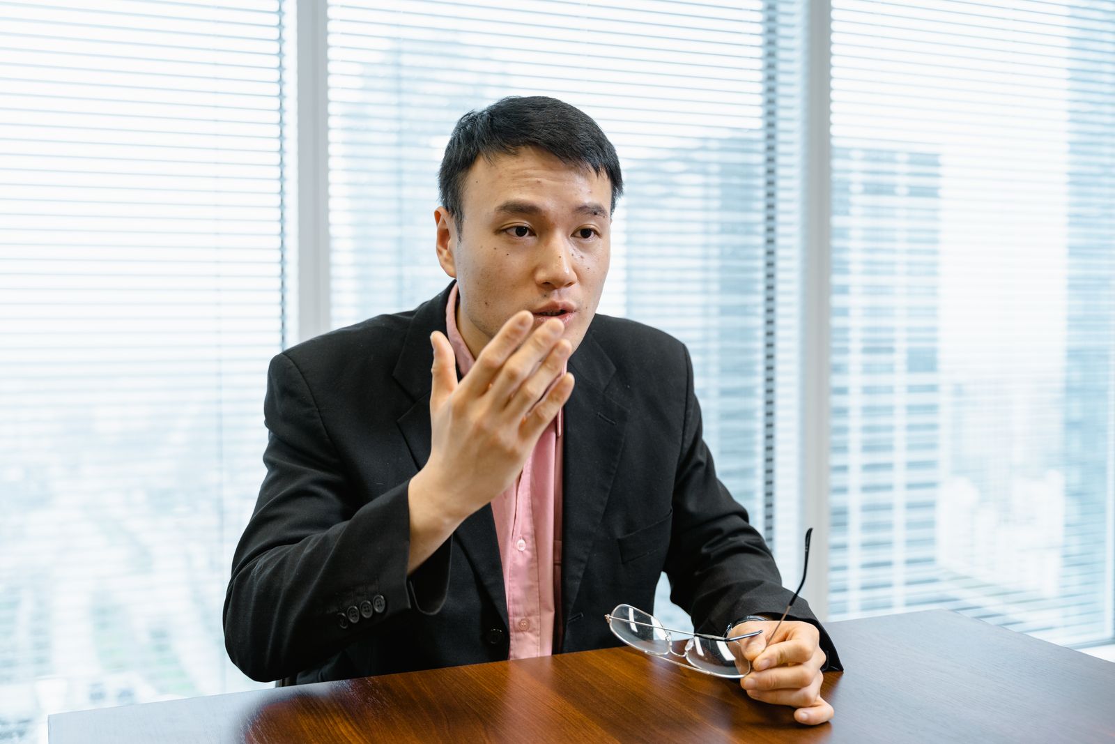 Man in a black blazer speaking in an office with a city view behind him — photo by MART PRODUCTION on Pexels