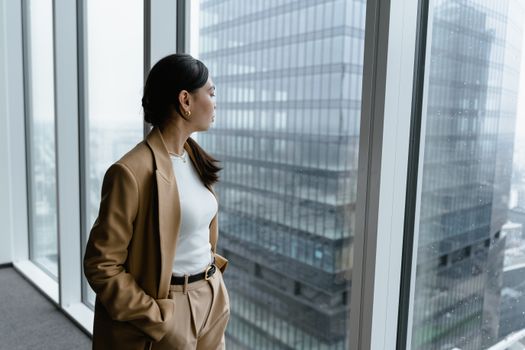 Businesswoman in brown coat looking out window in modern office.