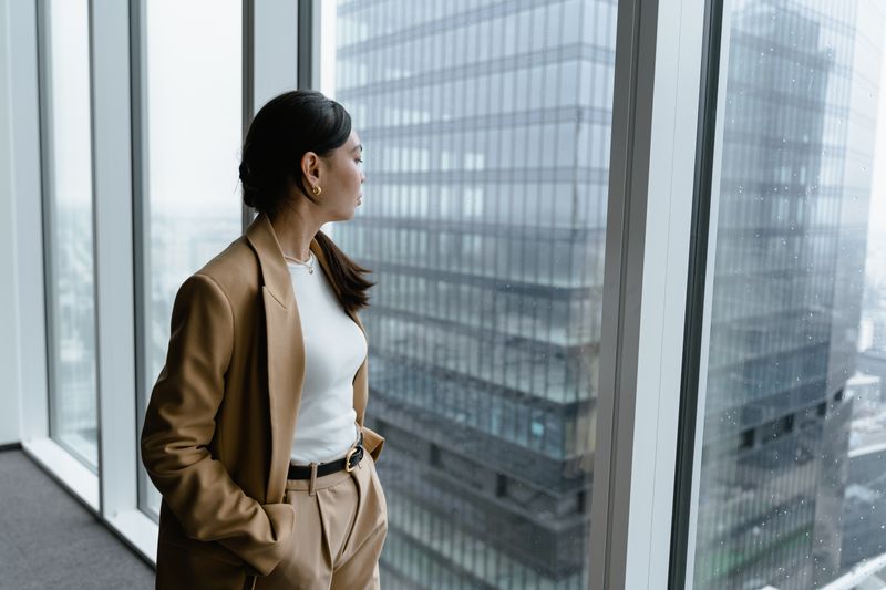Frau im Blazer blickt aus dem Fenster eines Büros im Hochhaus