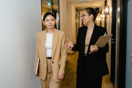 Two businesswomen converse while walking through a modern office hallway, holding documents.