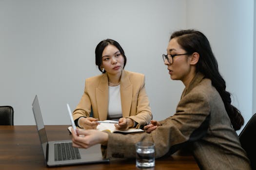 Two Asian businesswomen in a professional meeting discussing work at a conference table.