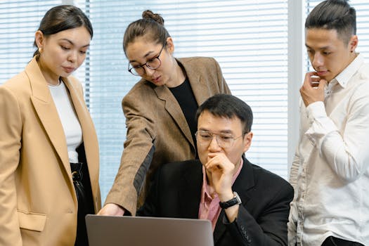 A group of focused professionals collaborating around a laptop in a modern office setting.