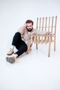 Trendy young bearded male in gumshoes and pants looking at camera while leaning with hand on wooden chair