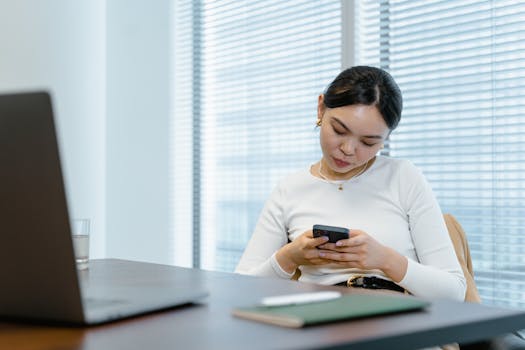 Asian woman sitting at her desk, texting on a smartphone in a modern office setting.