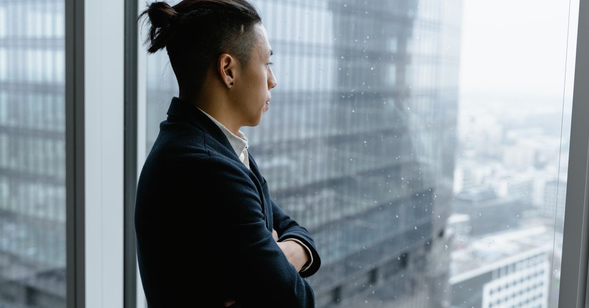 Asian businessman in formal attire looks out from office window on a rainy day.