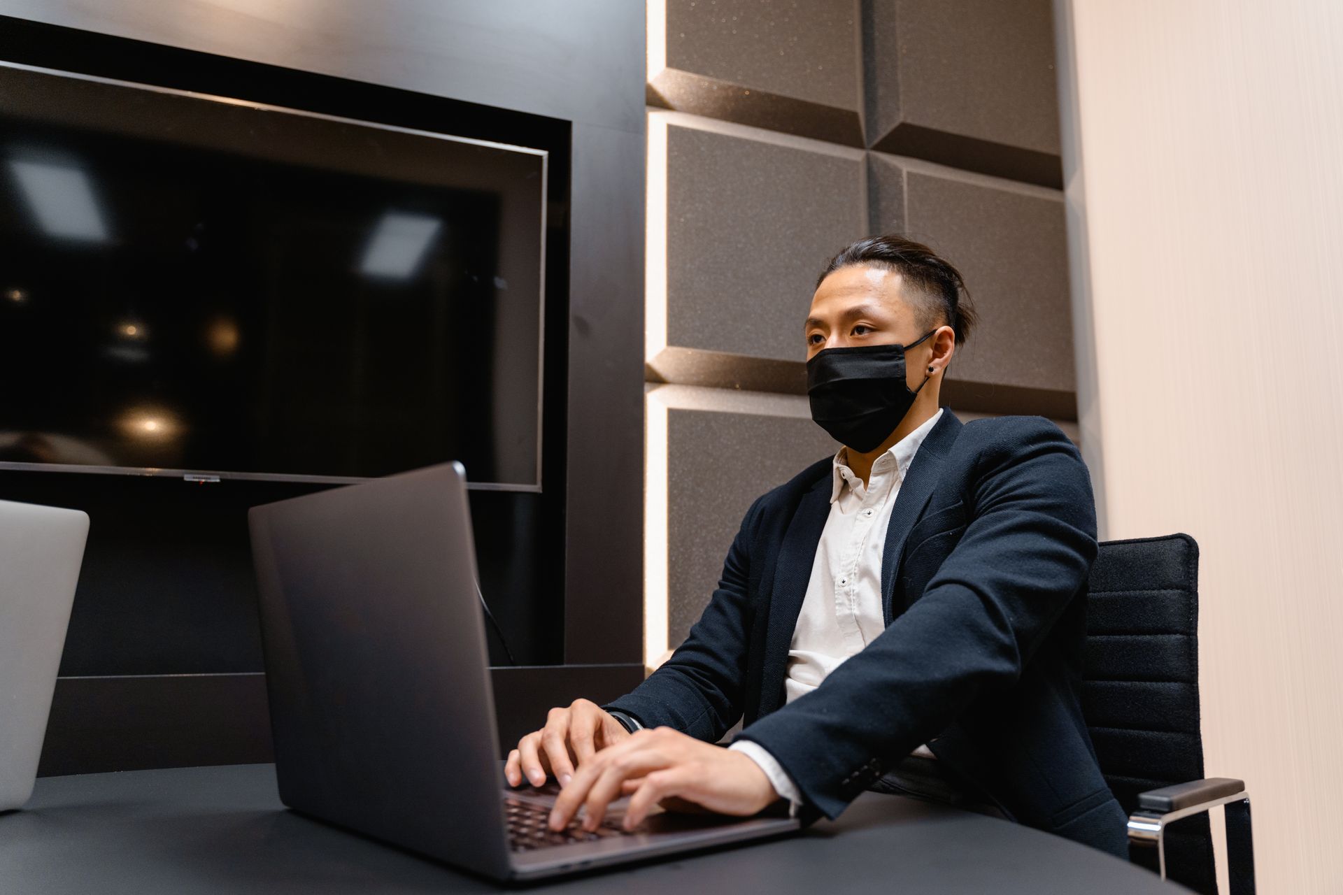 Asian man in face mask working on laptop in modern office setting.