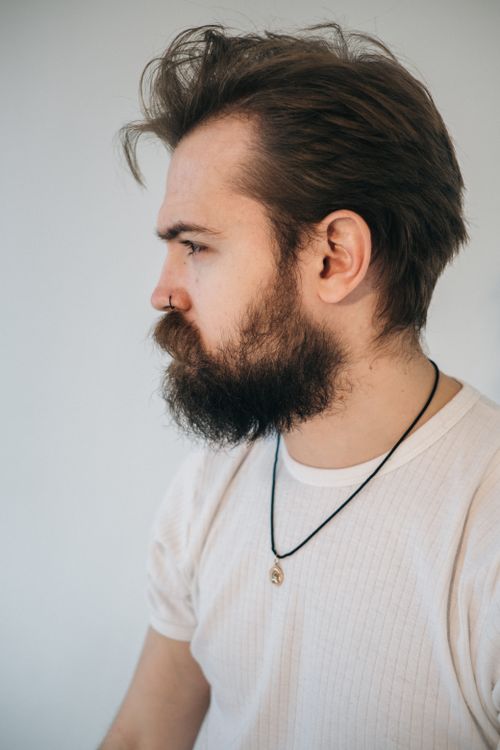 Free Young bearded male with brown hair in t shirt and pendant looking forward on light background Stock Photo